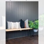 A modern room with a black vertical shiplap accent wall, a wooden bench with black metal legs, four decorative pillows, and a potted plant on the right. Text at top reads: “DIY Black Vertical Shiplap Accent Wall Tutorial.”.