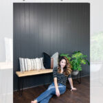 A woman sits on a dark wood floor in front of a black vertical shiplap accent wall. A bench with black legs, neutral pillows, and a green plant are placed against the wall. Text above reads: DIY black vertical shiplap accent wall tutorial.