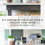 A rustic outdoor table with potted plants, a drink setup, a mini pizza oven, and a sign reading “It’s 5 o’clock somewhere,” sitting against a white siding wall.