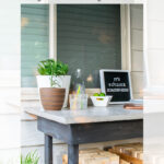 A modern outdoor bar table with a concrete tabletop, plants, drinks, and a sign reading “It’s 5 o’clock somewhere.” Wooden crates are stored underneath, with a house window and siding in the background.