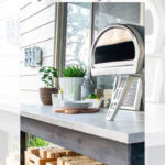 Outdoor kitchen scene featuring a DIY concrete tabletop on a wooden frame, set with plates, utensils, and potted herbs, with a pizza oven on top and stacked firewood underneath. Text overlay explains the DIY project.