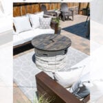 A modern patio features a round gas fire pit with a wooden cover, surrounded by white cushioned seating, a patterned rug, and brick pavers, creating a stylish outdoor lounge area.