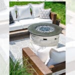 A modern outdoor patio features a circular gas fire pit with a stone base and wood cover, surrounded by white cushioned sectional seating and geometric-patterned pillows. Grass and pavers complete the cozy setting.
