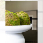 A white pedestal bowl holding two moss-covered decorative spheres sits on a dark table. The image is overlaid with text about a DIY Dollar Tree decorative pedestal bowl centerpiece by Garrison Street Design Studio.