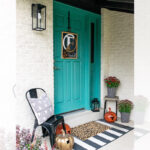 A turquoise front door is decorated for fall with a monogram wreath, striped doormat, metallic pumpkins, patterned planters with flowers, and a black chair with a geometric cushion. The porch has a cozy, modern vibe.