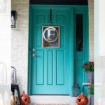 A teal front door decorated for fall with a monogram wreath, two carved pumpkins, potted mums, and autumn decor on a porch. A black chair and lantern are also placed near the door.
