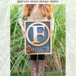 A woman in a patterned sweater and taupe boots stands on grass holding a square chalkboard wreath sign with a large letter "F," pinecones, and “Happy Fall” written in chalk. Tall green grass is in the background.