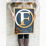 A person in a patterned sweater and tall brown boots holds a chalkboard wreath sign with a large gold letter "F," pinecones, and the handwritten words "Happy Fall" against a light brick wall.