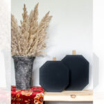 A marbled vase with tall dried pampas grass and two geometric black wooden pumpkins sit on a wooden shelf with a colorful floral fabric accent. Text above reads: "DIY Fall Scrap Wood Pumpkins, garrison street design studio.