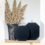 A decorative fall display featuring a marbled vase filled with beige pampas grass and two black, geometric wooden pumpkins with small wood stems, set on a light wooden shelf. Text reads "DIY Fall Scrap Wood Pumpkins.