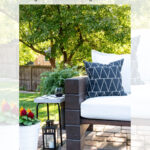 Outdoor patio scene with a white cushioned sofa, dark wood armrest, a black and white geometric pillow, a small black lantern, and potted flowers on a paved patio, with trees and a wooden fence in the background.