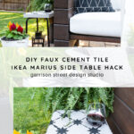 A small outdoor side table with a black and white faux cement tile top holds a glass of red wine. Next to it, a wicker chair with a black and white pillow sits on a patio, surrounded by green plants and flowers.