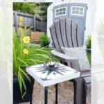 A small side table with a faux cement tile top sits next to a brown outdoor chair and a black-and-white pillow. Yellow flowers, green plants, and a gray shed are in the background. Text overlays the image.