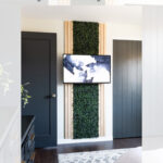 A modern living room features a faux green living wall behind a mounted TV, framed with wood panels. Dark doors, light walls, and a patterned rug complete the stylish, minimal decor.