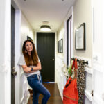 A woman stands smiling in a bright, narrow hallway with white ladder board and batten walls, black door at the end, and decor items including bags and a leopard-print scarf hanging on the right. Text overlay reads, "DIY Hallway Makeover with Ladder Board and Batten.