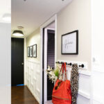 A modern hallway with white board and batten walls, framed art, a wall hook holding a red purse and leopard scarf, and a vase with white flowers on a small table. Text above reads, "DIY Hallway Makeover with Ladder Board and Batten.