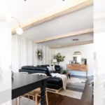 Bright living and dining area with black furniture, wood flooring, area rugs, and faux wood ceiling beams. Modern decor includes a sunburst mirror, plants, and minimalist white paneled walls. Text overlay explains a DIY beam project.