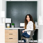 A woman with long brown hair sits at a white and wood fluted desk against a dark green wall, smiling at the camera. Text above reads, "DIY Ikea Rast Fluted Dresser Desk Hack - Garrison Street Design Studio.