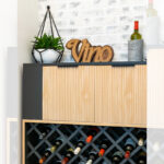 A modern DIY wine bar cabinet with a wooden slat front, black shelf, and lattice wine rack filled with bottles. On top are a potted plant, a “Vino” sign, and wine bottles, set against a white brick wall.