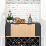 A modern wine bar cabinet with a geometric wine rack holding bottles, a wood-paneled drawer, a plant, a "Vino" sign, a wine bottle, and glasses on top, styled against a white brick wall.