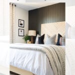 A modern boho bedroom with a chevron wood headboard, white bedding with black accents, geometric pillows, and a patterned rug. The wall is painted dark, with neutral decor and a lamp on the nightstand. Text reads: "DIY Modern Boho Bedroom Makeover Reveal.