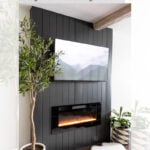 A modern living room features a black shiplap accent wall with a mounted TV above an electric fireplace. Green plants and a white rug add cozy, contemporary decor to the space. Text overlay reads “Modern DIY Electric Fireplace and Shiplap TV Wall.”.