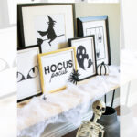 A Halloween-themed display features framed black-and-white prints with spooky designs, surrounded by faux spider webs, spiders, and a skeleton sitting on a bench.