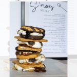 A stack of four s’mores with toasted marshmallows and melted chocolate on a wooden board, in front of a printed s’mores menu and modern decor background.