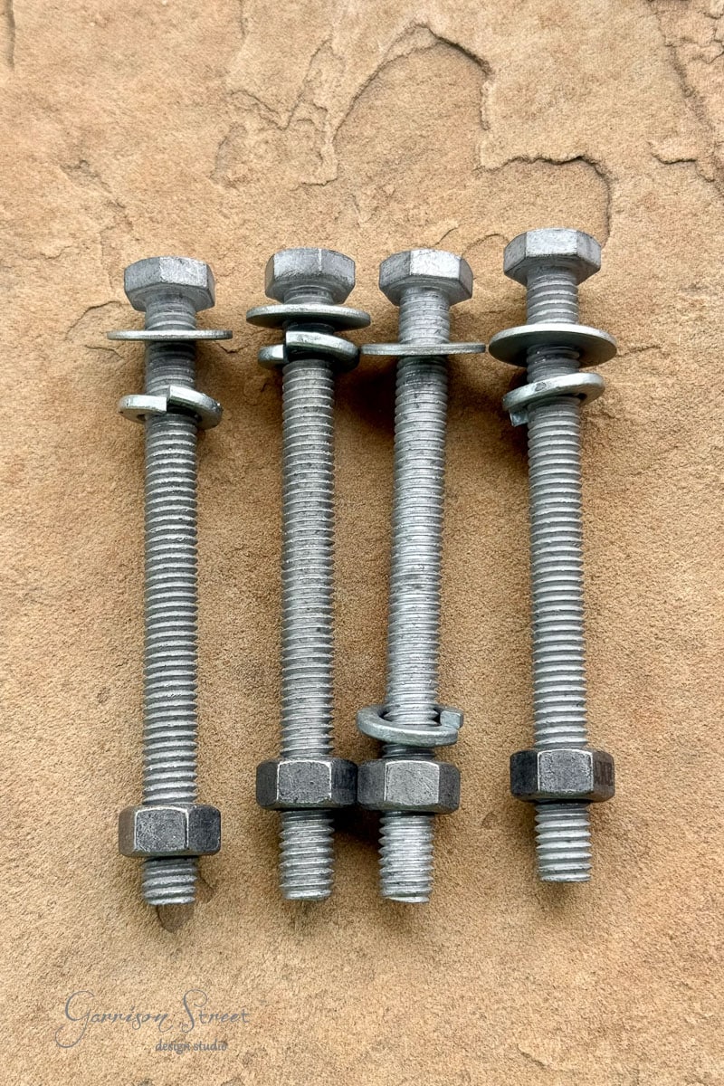 Four metal bolts with hex nuts and washers are aligned vertically on a textured, tan surface. The bolts are arranged side by side, each threaded with two washers and two locknuts.