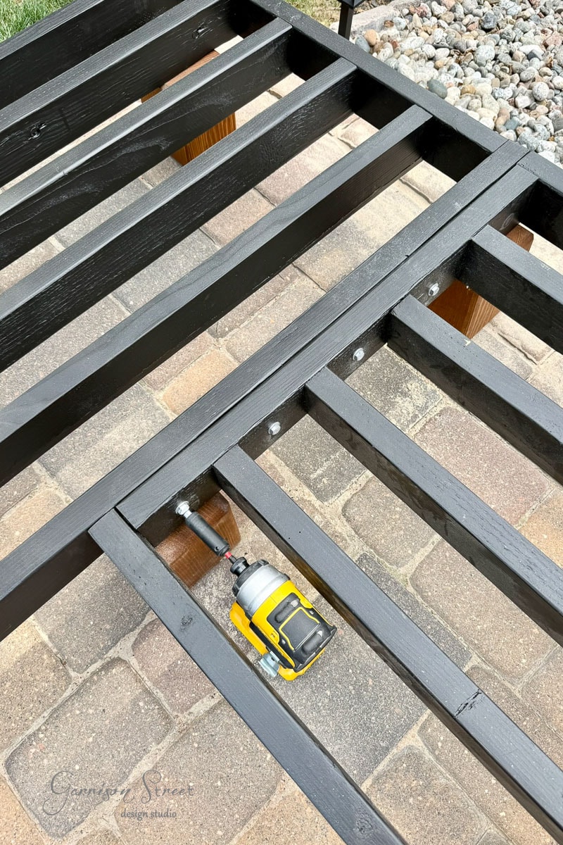 A yellow power drill rests on a black wooden frame being assembled on a patio made of stone pavers; bolts connect the frame pieces, with rocks visible in the background.
