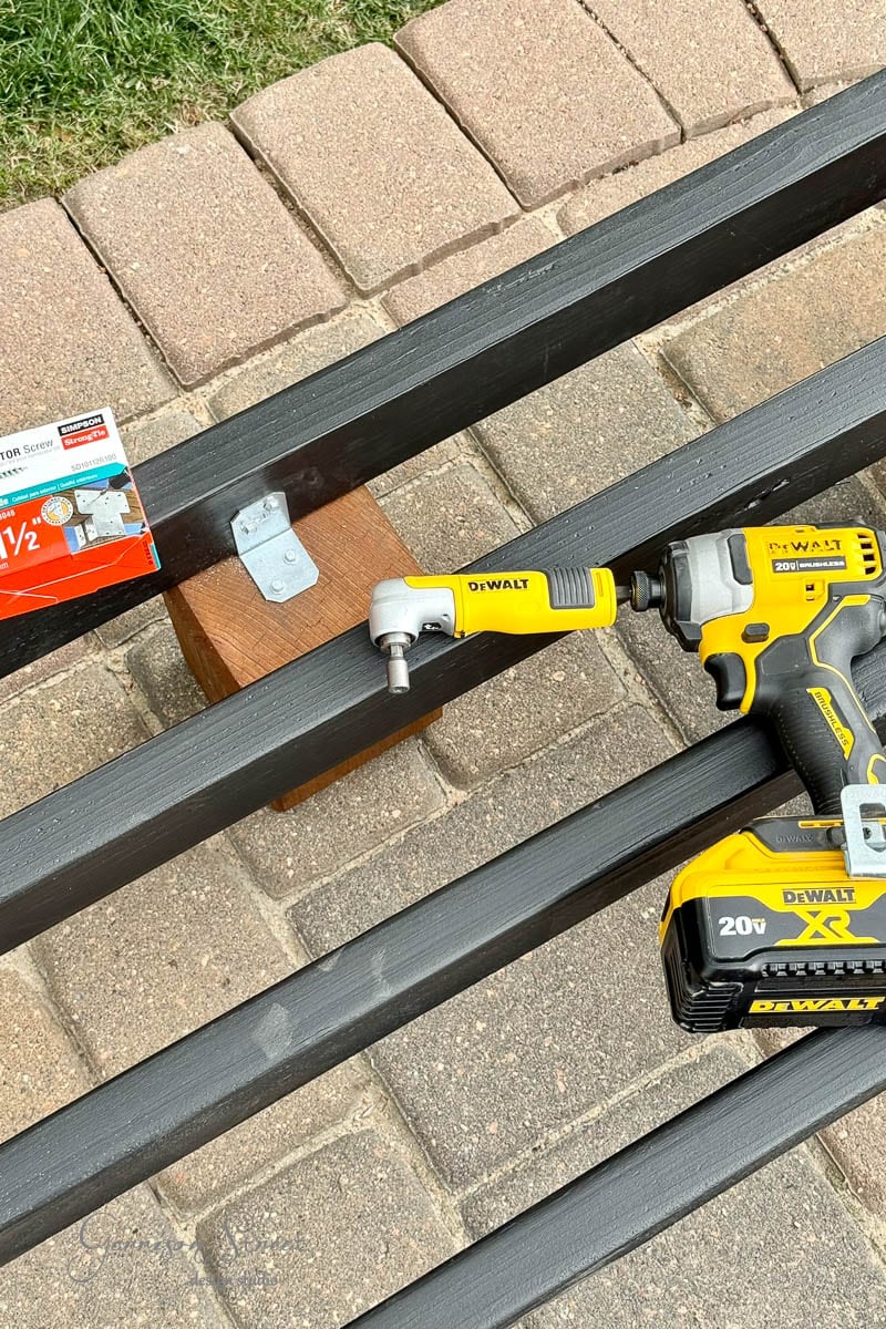 A DeWalt power drill with an attachment rests on black 2x4 frame near a bracket and a box of screws. The scene is outdoors, with brick paving and grass visible in the background.
