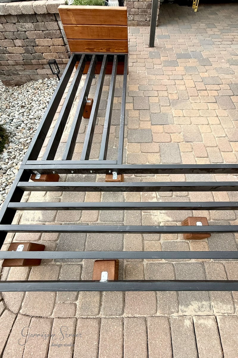A wood sectional frame sits on a stone patio, viewed from above. The frame rests on wooden blocks, with a wooden 6x6s attached. Rocks and bricks line the edges of the patio.