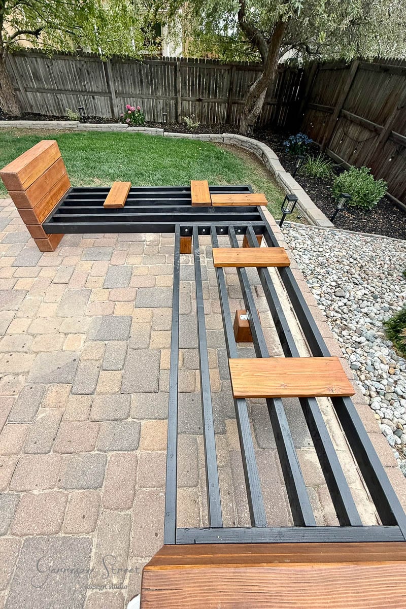 A wood outdoor bench frame is positioned on a stone patio, with some wooden slats attached as seats and backs. The frame is L-shaped and set near a landscaped garden with trees, rocks, and grass in the background.