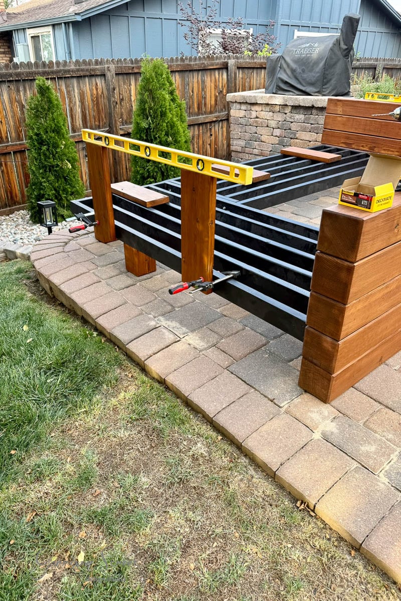 A partially built outdoor sectional with wood framing, a yellow level placed on top, clamps securing beams, and tools nearby. The furniture sits on a paved patio, with grass and a wooden fence in the background.
