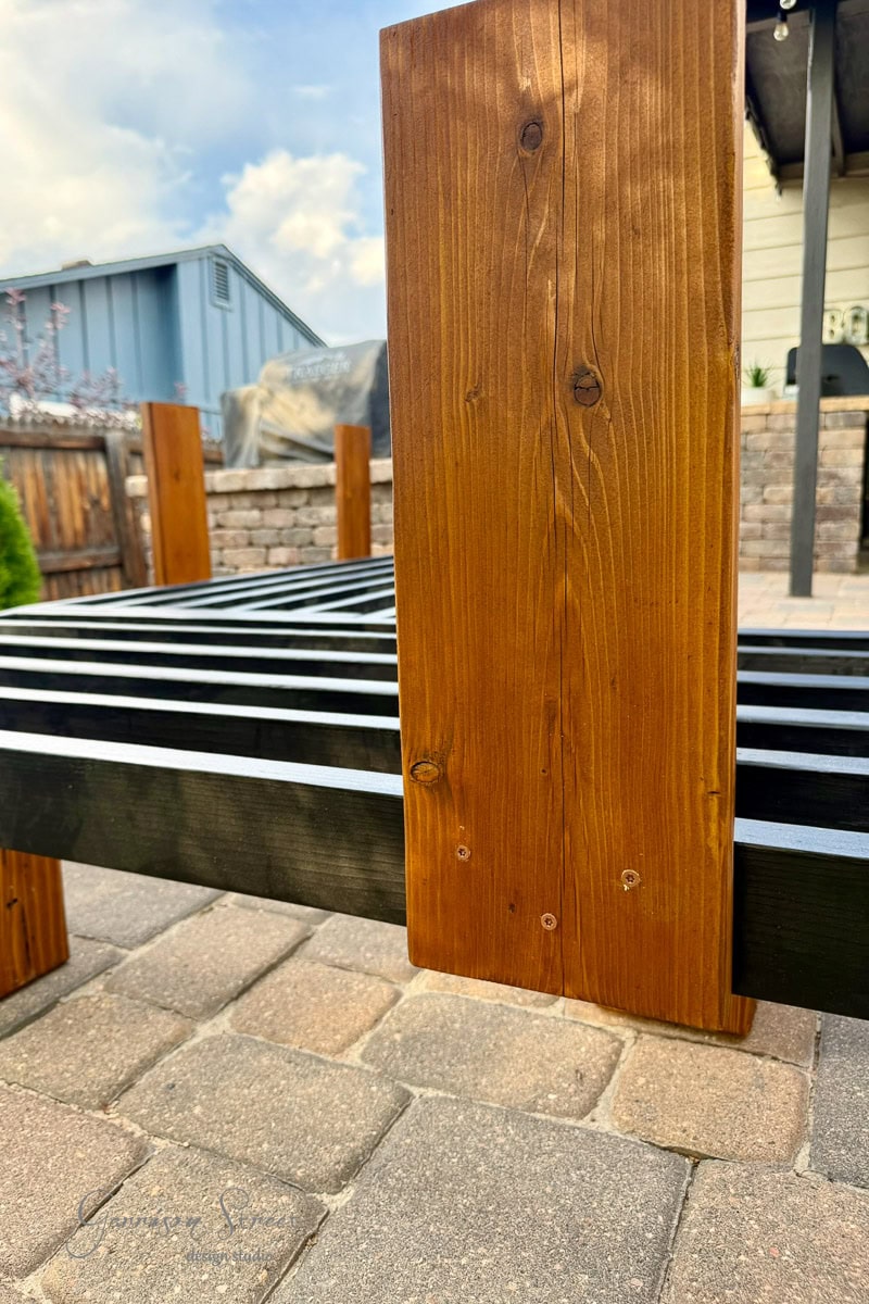 A close-up of a wooden outdoor bench being assembled on a stone patio, showing vertical and horizontal wooden slats, with houses and a cloudy sky in the background.