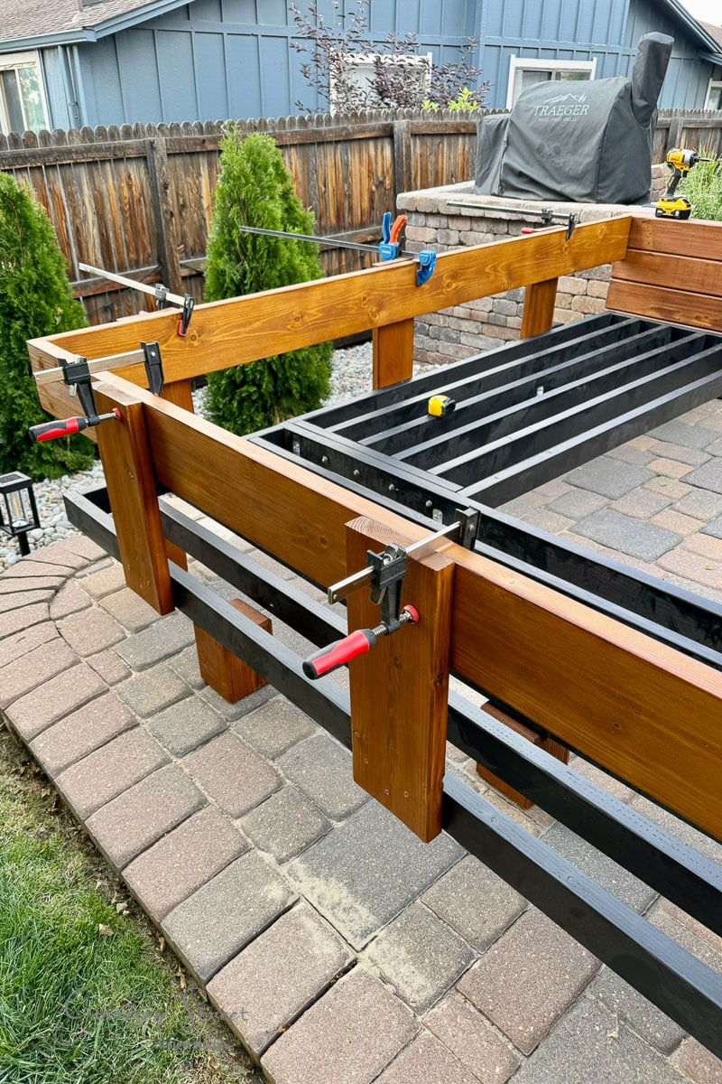 A wooden frame is being assembled with clamps holding pieces in place. The structure sits on a brick patio near a backyard fence, with tools and greenery visible in the background.