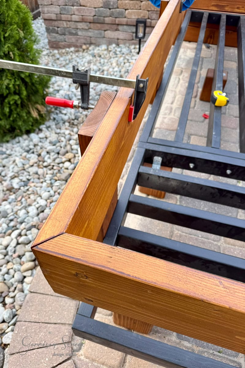 A close-up of a wooden frame being clamped together outdoors, with rocks and a brick wall in the background. A yellow tape measure rests on the black frame.