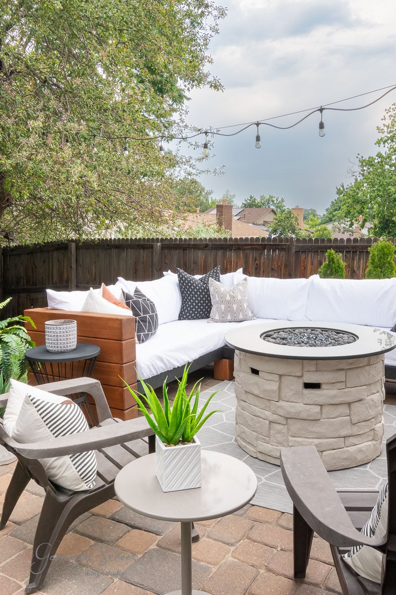 Cozy patio with a white cushioned sectional, black and white pillows, fire pit, chairs, potted plant on a small table, string lights overhead, and greenery in the background.