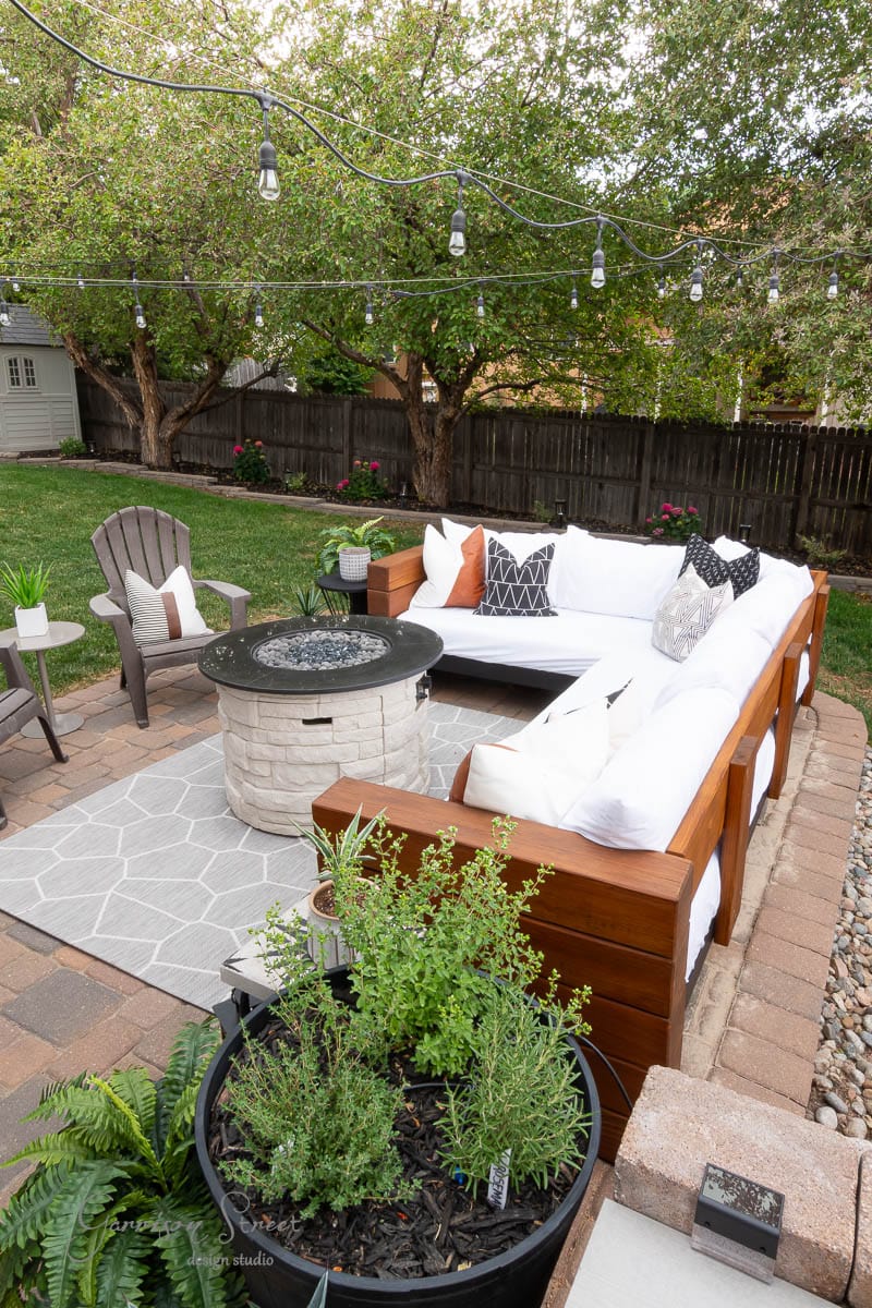 A cozy backyard patio with a white sectional sofa, gray chairs, a round fire pit, string lights, a patterned rug, and a planter with herbs, surrounded by trees and a wooden fence.