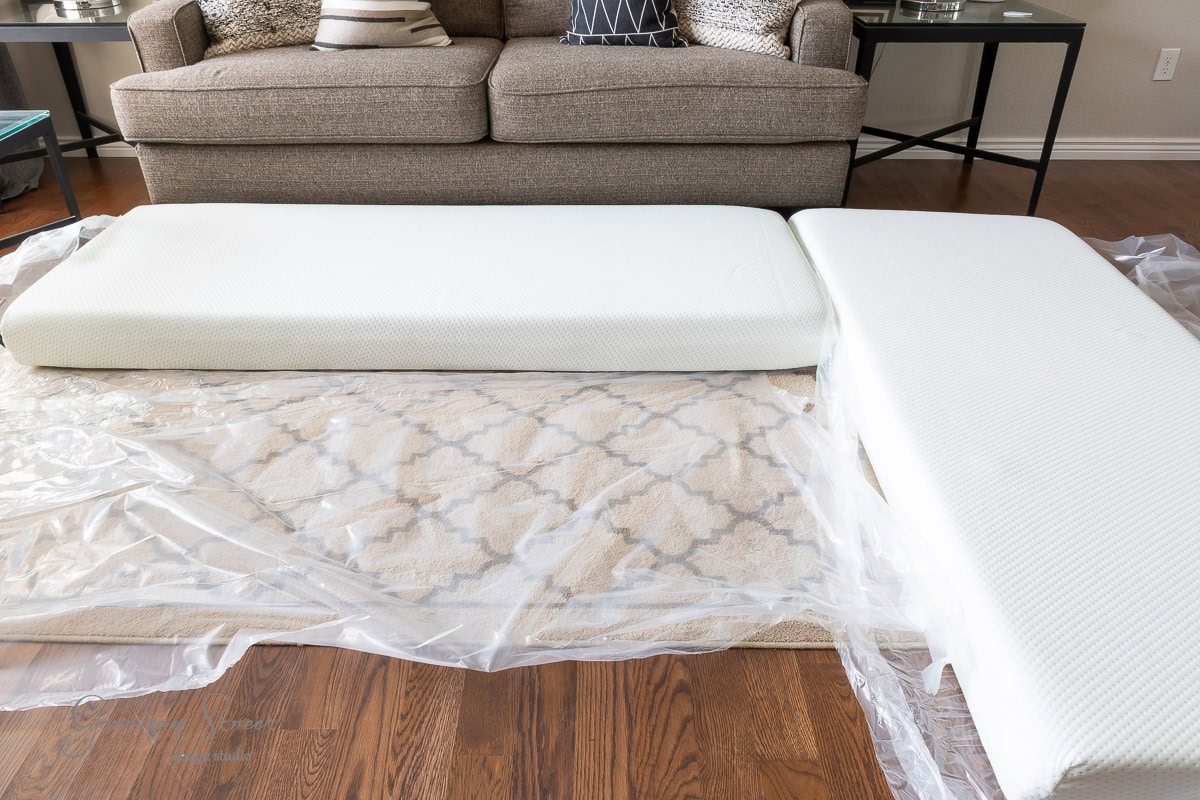 Two memory foam mattresses are unrolled on a clear plastic sheet over a patterned rug and wooden floor in front of a beige couch.