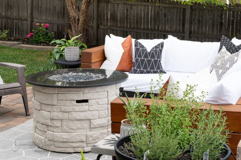 Outdoor patio with a white stone fire pit, a white cushioned sectional sofa with patterned pillows, green plants, and a wooden fence in the background.