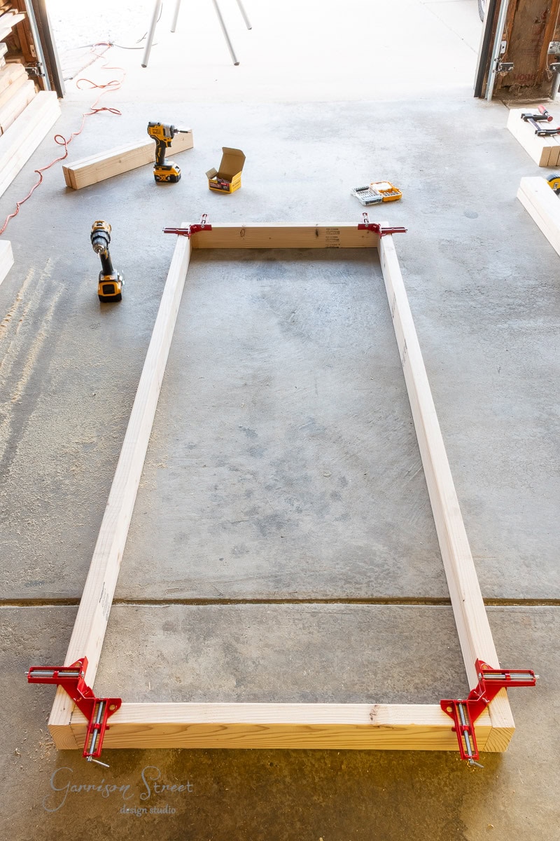 A rectangular wooden frame is clamped at the corners on a concrete floor. Power tools, screws, and other wooden pieces are nearby, suggesting a woodworking project in progress.