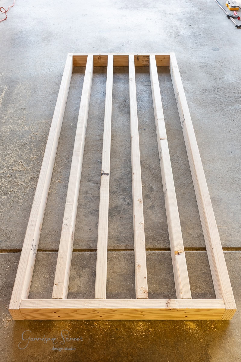 A wooden frame made of light-colored lumber lies on a concrete floor. The frame consists of two long side pieces, two shorter end pieces, and five evenly spaced vertical crossbars. Tools are partially visible in the background.