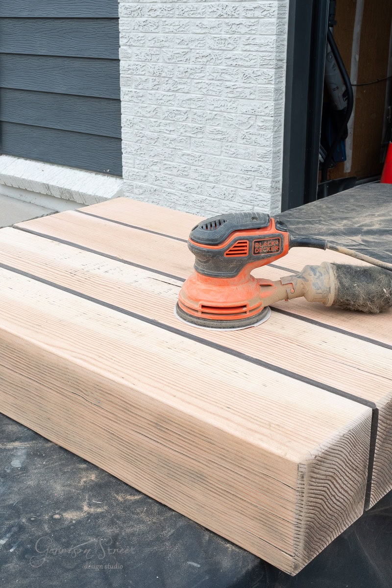 An orange electric sander rests on top of large, unfinished 6x6 wooden blocks, which are placed outdoors near a white brick wall and a partially open garage.
