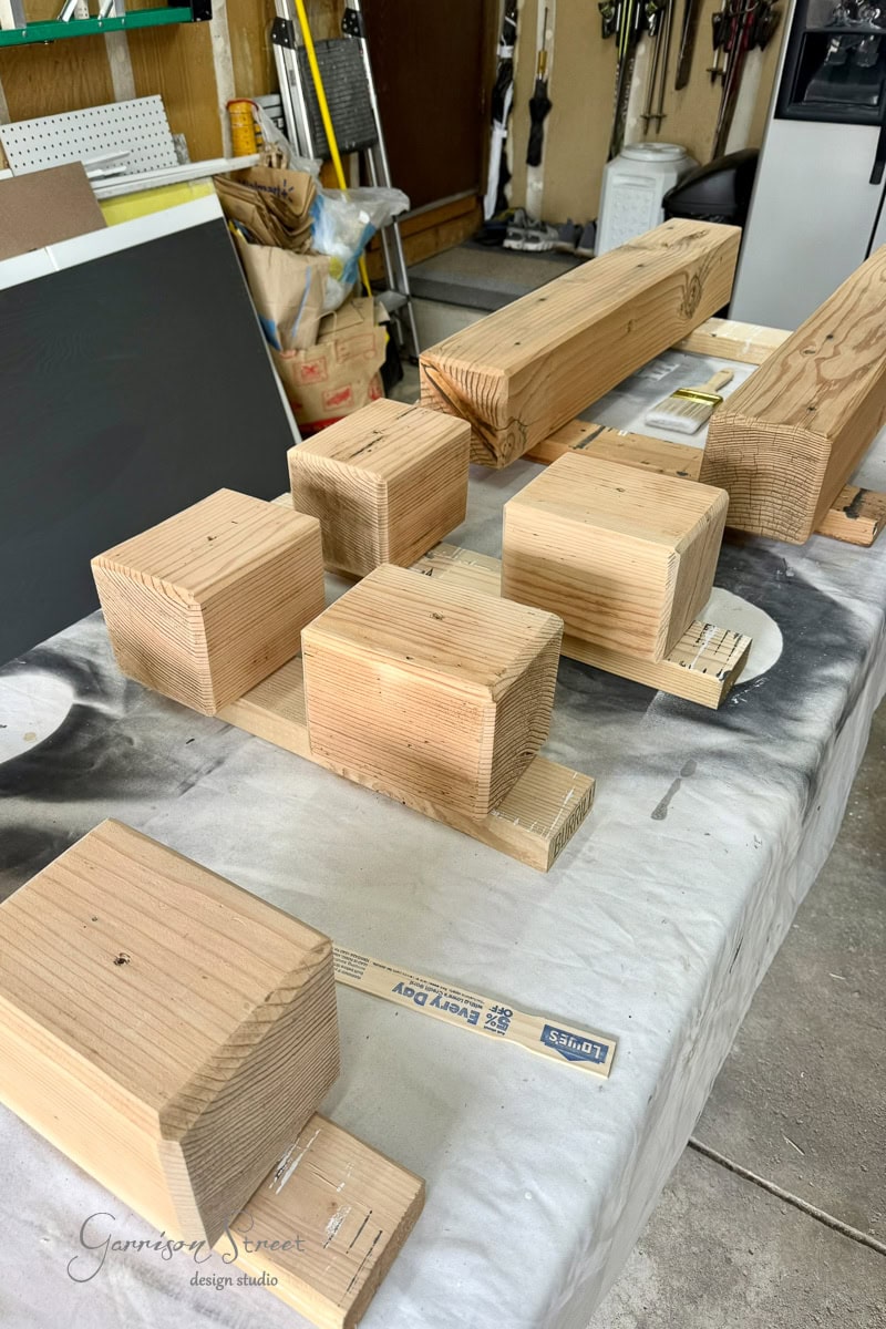 Several wooden 6x6 blocks of different sizes are arranged on a table in a workshop, ready for a woodworking project. Tools and supplies are visible in the background.
