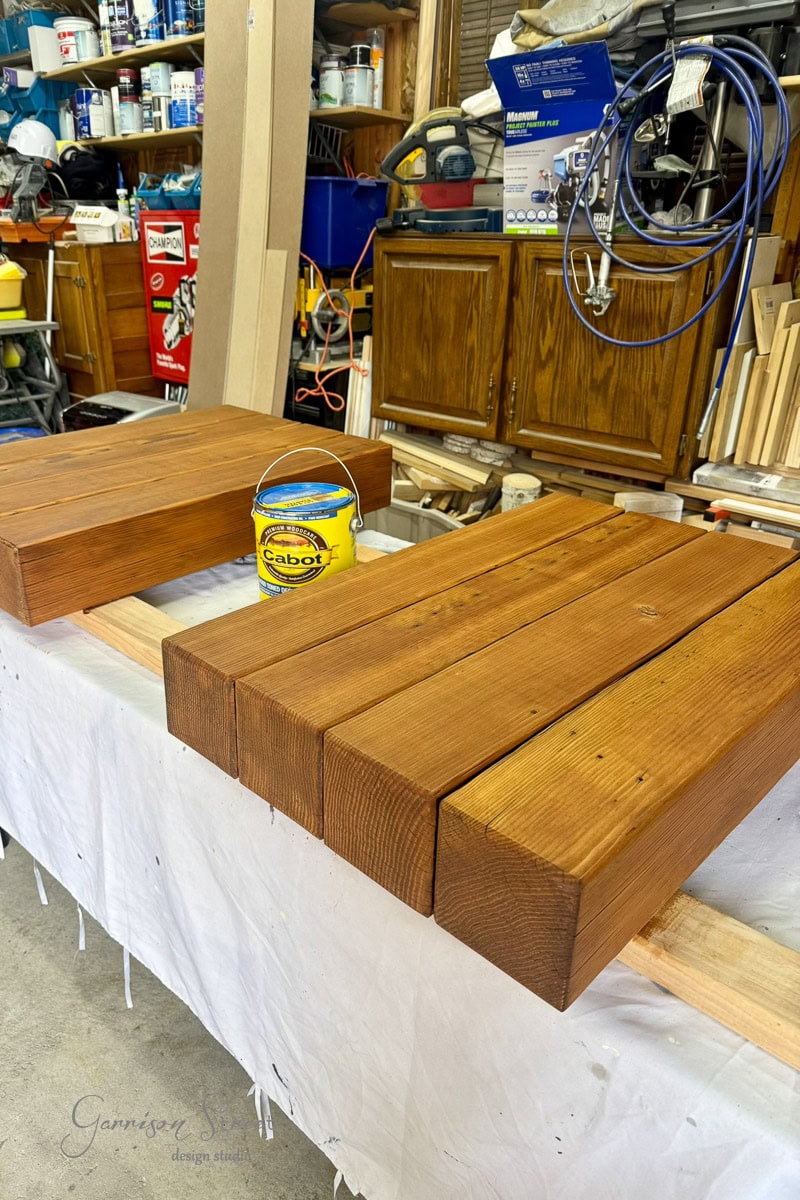 Two stained wooden stacks of 6x6s drying on a covered table in a cluttered workshop, with a yellow can of Cabot wood finish nearby and various tools and supplies in the background.