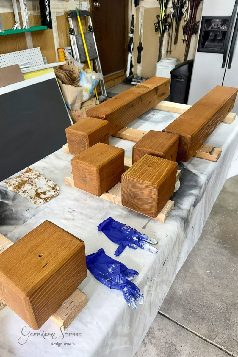 Wooden blocks and beams are drying on a makeshift table in a garage workshop. Two blue paint-stained gloves lie nearby on a white cloth. Tools and equipment are visible in the background.