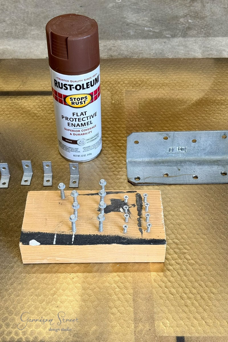A can of brown Rust-Oleum spray paint, metal brackets, a painted wooden block with screws, and are arranged on a gold-textured surface.