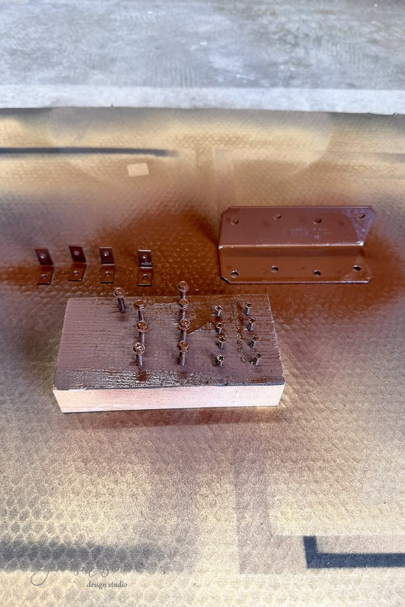 Metal hardware pieces, screws, and brackets are arranged on a surface and a wooden block, all spray-painted brown, with the surrounding area covered in overspray.