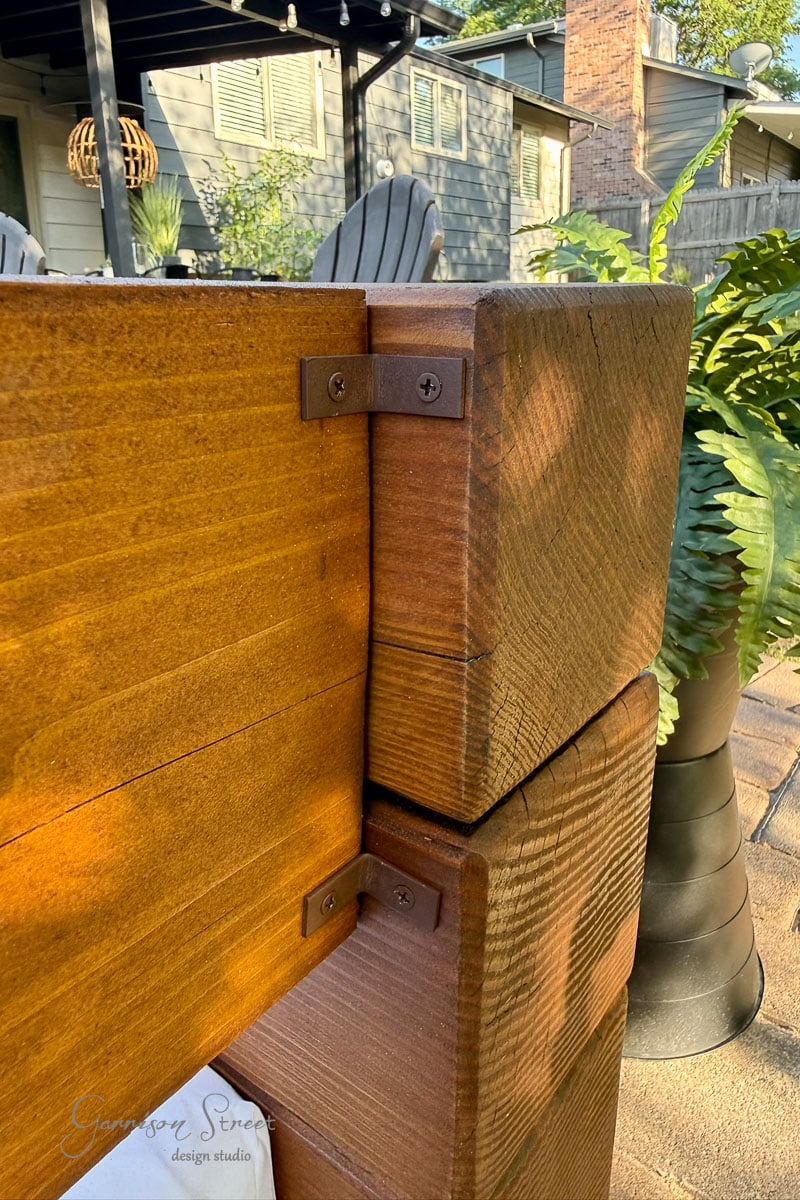 Close-up of two large wooden 6x6s joined with a 2x8 by metal angle brackets on a patio, with a potted fern and patio chairs visible in the background.